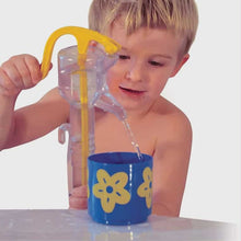Child pouring water from a yellow and clear container into a blue cup with yellow flowers on a white background