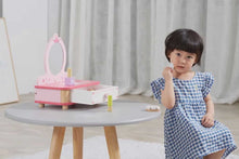 Child in a blue checkered dress sitting next to a pink toy vanity set on a gray table.