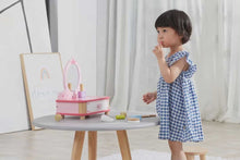 Child playing with a pink toy vanity set on a small table in a room with white curtains.